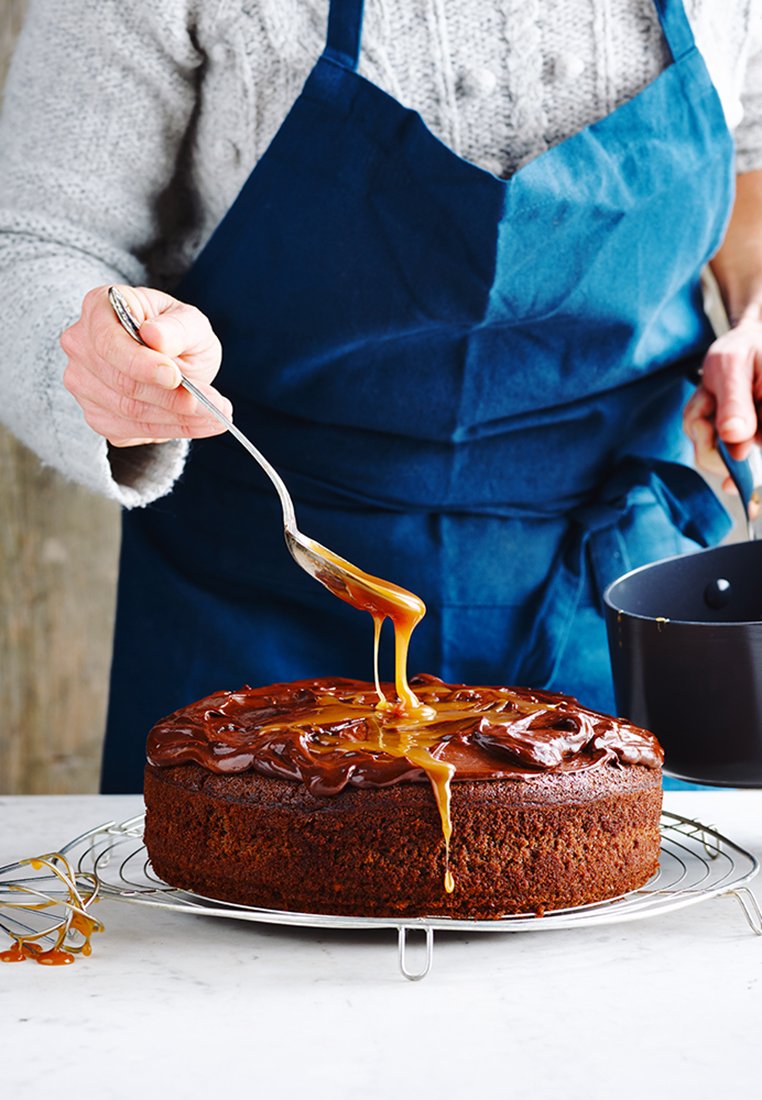 Chocoladetaart op een metalen koelende rek, bedekt met glanzende karamel die eroverheen druppelt. Een persoon in een blauwe schort giet karamel uit een zwarte pan.