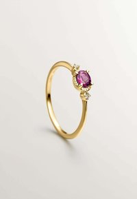 Anillo de oro con una piedra preciosa ovalada de color rosa en el centro, sujeta por cuatro garras, flanqueada por dos pequeñas piedras claras a cada lado. Diseño de banda lisa.