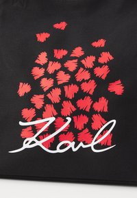 Sac fourre-tout noir avec un motif de cœurs en coups de pinceau rouges et une signature blanche "Karl" en bas, fabriqué en matériau durable.