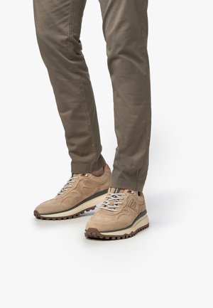 Beige suède sneakers met een lichte zool, textuuraccenten en grijze veters, gedragen met lichtbruine slim-fit broeken.