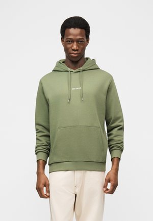 Moški v maslinasto zeleni kapucarji z žepom spredaj in vrvicami, stoji pred enobarvnim svetlim ozadjem.