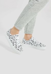 Baskets blanches avec un motif de chien en bande dessinée noir, bout rond, semelle plate et design classique à lacets, associées à un pantalon de survêtement gris.