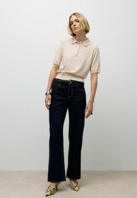 Hellbeige Poloshirt mit kurzen Ärmeln, kombiniert mit dunkelblauen Schlagjeans. Accessoires mit einem braunen Gürtel und Schlangenleder-Absätzen.