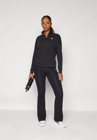 Svart atletisk outfit med en zip-nek pullover och figurnära byxor. Tillverkad av ett mjukt tyg med en subtil lyster, accentuerad med en logotyp.