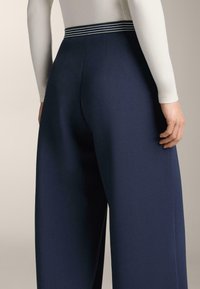 Osoba oblečená v námorníckych širokých nohaviciach s pruhovaným pásom a priliehavým bielym topom s dlhým rukávom, zobrazená zo zadnej strany od pásu dolu.