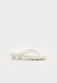 White thong sandal with low block heel and textured sole, featuring a cushioned strap and gold logo detail on a plain background.