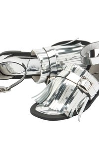 Metaliczne srebrne sandały z frędzlami, regulowanym paskiem w okolicy kostki i czarną podeszwą. Gładka i odbijająca powierzchnia z detalem w postaci klamry.