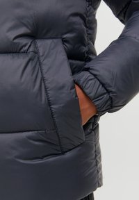 Temno moder puhovka z gladkim, sijajnim zaključkom. Vključuje veliko, kvadratno žep in elastične rokave, kar poudarja toploto in udobje.