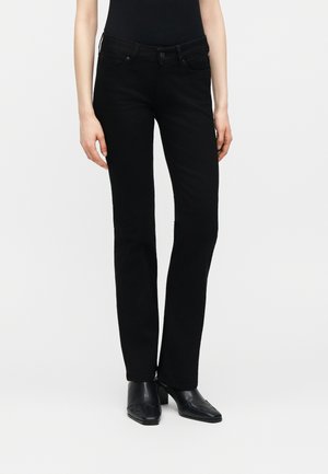 Γυναίκα που φοράει μαύρο τζιν με ίσια γραμμή, μαύρο εφαρμοστό τοπ και μαύρα μυτερά μποτάκια με τακούνι, στέκεται μπροστά σε λευκό φόντο.