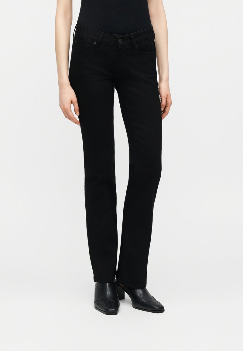 Γυναίκα που φοράει μαύρο τζιν με ίσια γραμμή, μαύρο εφαρμοστό τοπ και μαύρα μυτερά μποτάκια με τακούνι, στέκεται μπροστά σε λευκό φόντο.