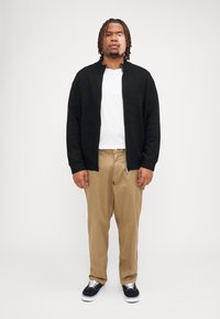 Sort zip-up sweatshirt, hvid t-shirt, brune bukser og sorte sneakers. Sweatshirten har ribbet krave og ærmer. Glat stofstruktur.