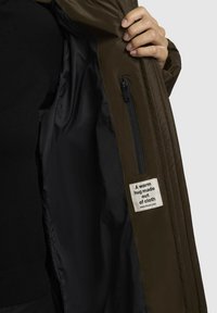 Olivovo zelená bunda s hladkým povrchom, ktorá má čiernu podšívku, zipsovú kapsu a štítok s nápisom "Teplé objatie z látky."