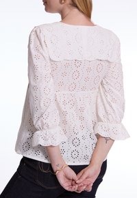 Witte broderie anglaise blouse met een gegolfde kraag, puffed mouwen en gerimpelde manchetten. Heeft doorlopende bloemen uitsnijdingen.