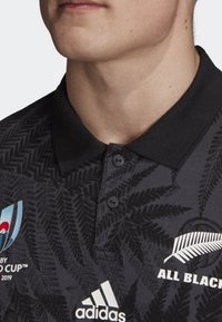 Svart pikétröja med krage, med ett mönster av ormbunkar, två knappar och vita logotyper för Rugby World Cup och Adidas.