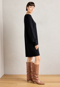 Robe noire tricotée avec une coupe ample, des manches longues et un col arrondi, assortie à des bottes en cuir marron texturé jusqu'aux genoux.
