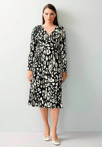 Schwarzes Kleid mit weißem abstraktem Muster, langen Ärmeln und V-Ausschnitt, aus einem glatten Stoff, knielang, kombiniert mit weißen Absätzen.