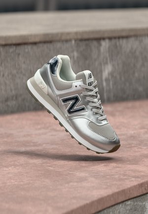Scarpa New Balance argento e grigia nell'aria sopra gradini in pietra con suola in gomma texturizzata e lacci e logo visibili.