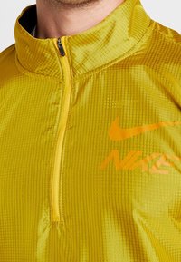 Chaqueta amarilla de Nike con tejido texturizado, parte delantera con media cremallera, cuello alto y logo naranja en contraste impreso en el pecho.