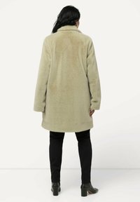 Cappotto lungo in morbido fleece beige con colletto alto, maniche lunghe e vestibilità comoda, abbinato a pantaloni neri aderenti e stivali con tacco.