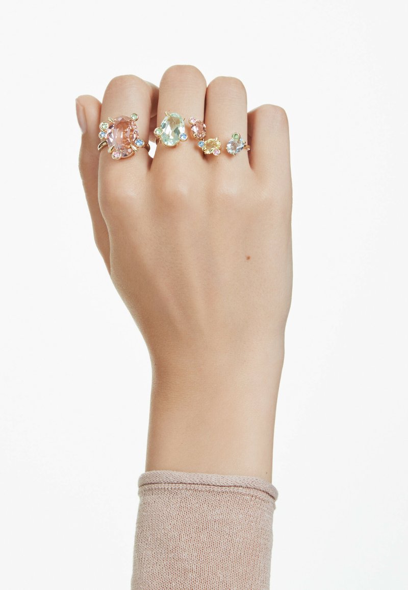 Fire ringe præsenteret på en hånd: en stor pink sten, to mindre pastelsten og et design med blandede farver, alle med guldfinish.