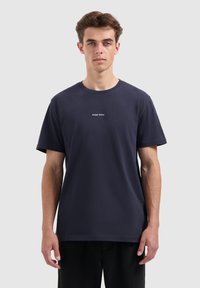 Jonge man in een marineblauwe T-shirt met de tekst "PURE PATH" op de borst, staande tegen een effen lichtgrijze achtergrond.