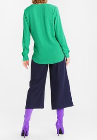 Vihreä pitkähihainen paita, jossa on pyöristetty helma, yhdistettynä tummansinisiin leveälahkeisiin housuihin ja kiiltoviininpunaisiin korkokenkiin. Silkkinen kangas, istuva malli.