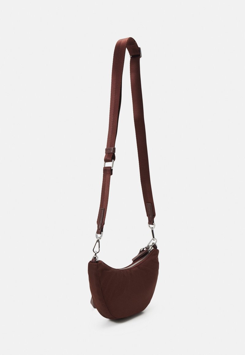 Bruine stoffen crossbodytas met een gebogen vorm, verstelbare band en zilveren hardware. Gladde textuur met een ritssluiting.