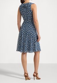Kleid mit Blumenmuster aus tiefblauem Stoff, ärmelloses Design, ausgestellter Rock, mit rüschem Ausschnitt und braunen High-Heels.