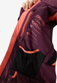 Giacca viola con accenti arancioni, realizzata in materiale impermeabile e con fodera in rete. Include etichette per le caratteristiche di prestazione e i dettagli dell'isolamento.