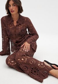 Brązowy zestaw na szydełku składający się z koszuli na guziki i pasujących spodni z kwiatowymi wzorami i wycięciami. Teksturowany materiał o otwartym splocie.