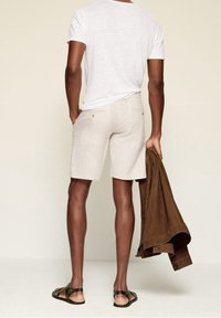 Weißes T-Shirt mit kurzen Ärmeln, hellbeige Shorts, braune Wildlederjacke in der Hand, schwarze Sandalen, von hinten betrachtet. Glattes, lässiges Design.