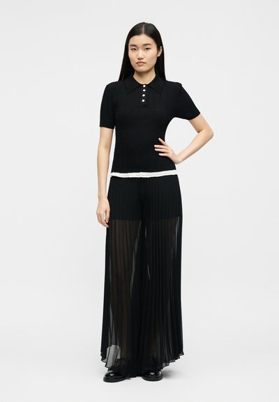 Γυναίκα που στέκεται με το χέρι στη μέση, φορώντας μαύρο κοντομάνικο τοπ με λευκή ρίγα και διαφανές μαύρο παντελόνι με φαρδιά μπατζάκια.