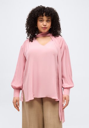 Žena s kudrnatými vlasy, která má na sobě volnou růžovou halenku s dlouhými rukávy a detailem šátku a béžové kalhoty s širokými nohavicemi, na jednobarevném pozadí.