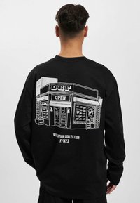 Sort langærmet sweatshirt med en hvid linjetegning af en butik, hvor teksten "DEF OPEN" og forskellige genstande i vinduet ses.