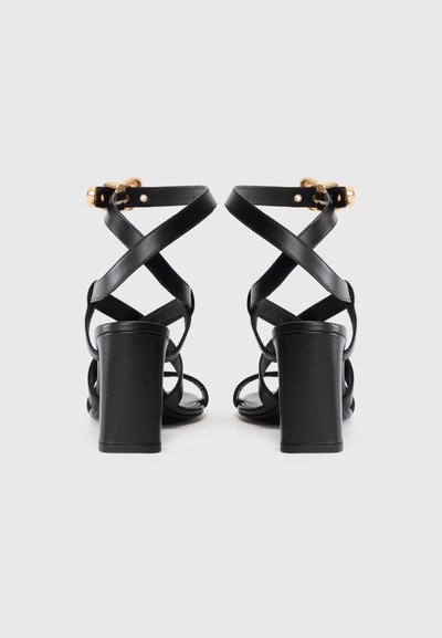 Een paar zwarte leren hoge hak sandalen met gekruiste banden en gouden gespsluitingen, gefotografeerd vanuit de achterkant op een witte achtergrond.