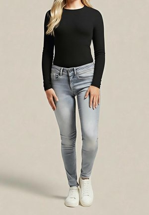 Vrouw die een zwarte top met lange mouwen, lichtgrijze strakke jeans en witte sneakers draagt, staand tegen een effen achtergrond.