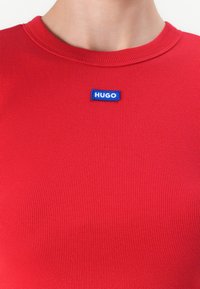 Raudonas raukšlėtas švarkas su apvaliu kaklu ir maža mėlyna "HUGO" logotipo etikete kairėje pusėje. Minkšta tekstūra, prigludęs dizainas.