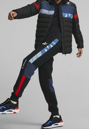 Person trägt schwarze Puma BMW Motorsport Jacke und Hose mit blauen, roten und weißen Akzenten, hebt ein Bein an und steht vor grauem Hintergrund.