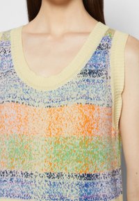 Femme portant un haut tricoté sans manches avec un motif multicolore comprenant des bandes horizontales jaunes, orange, vertes, bleues et violettes.
