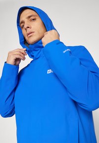 Veste de pluie bleue avec capuche, fabriquée en matériau lisse et résistant à l'eau. Présente un logo sur la poitrine et des poignets ajustables.