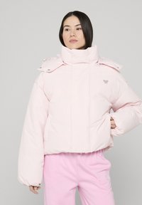 Vrouw draagt een lichtroze donsjack met hoge kraag en bijpassende roze broek, staand tegen een effen lichte achtergrond.