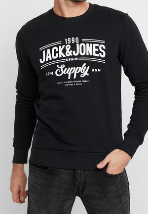 Czarna bluza z długim rękawem z białym napisem "1990 Jack & Jones Denim Supply", noszona przez osobę z jedną ręką w kieszeni, a drugą opadającą w dół.