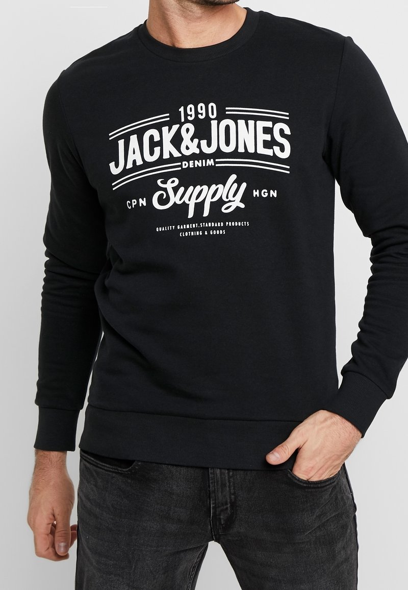 Sweat-shirt noir à manches longues avec texte blanc "1990 Jack & Jones Denim Supply" porté par une personne avec une main dans la poche et l'autre pendante.