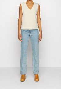 Beige gestrickter ärmelloser Pullover mit V-Ausschnitt, der ein strukturiertes Muster aufweist, kombiniert mit hellblauen geraden Jeans und braunen Stiefeletten.