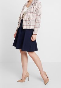 Rutig kort jacka i kräm, röd och marinblå, parat med en marinblå utsvängd kjol och nude högklackade skor. Guldknappar pryder jackan.