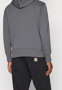 Šedá mikina s kapucí vyrobená z měkké látky, s podšívkou v kapuci, žebrovanými manžetami a lemem. Černé kalhoty s viditelnou nášivkou loga na zadní straně.