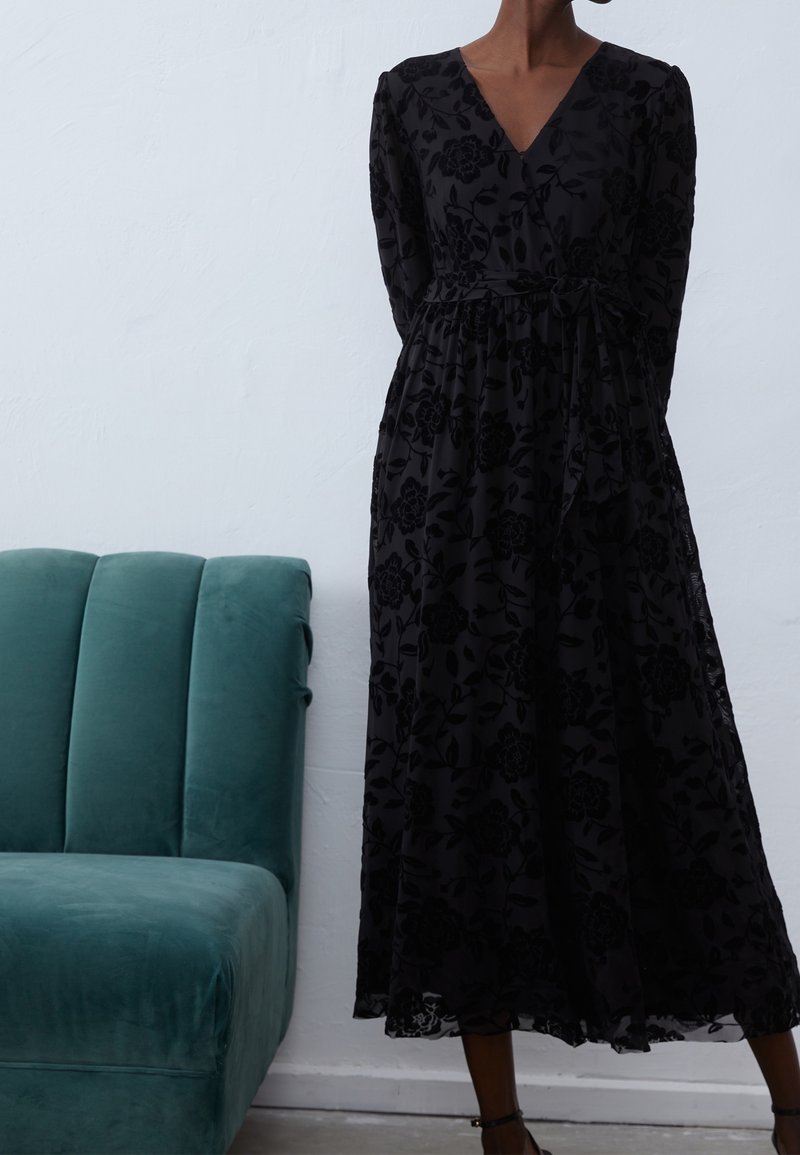 Czarna długa sukienka w kwiaty z długimi rękawami i dekoltem w kształcie litery V. Miękki, lejący się materiał z delikatnym wzorem, wiązana w talii.