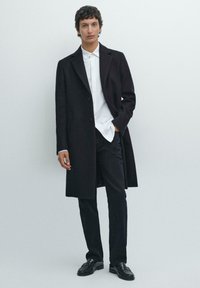 Cappotto di lana nero con un design sartoriale, chiusura a un bottone e tasche laterali, indossato sopra una camicia bianca e pantaloni neri con scarpe in pelle.