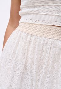 Jupe en dentelle blanche avec une taille élastique texturée. La dentelle présente des motifs complexes et un design doux et fluide.