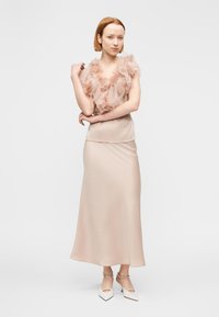 Robe fluide rose blush avec un décolleté en tulle fleuri et une jupe en satin. Le modèle l'associe à des chaussures blanches à bouts pointus et un bracelet.